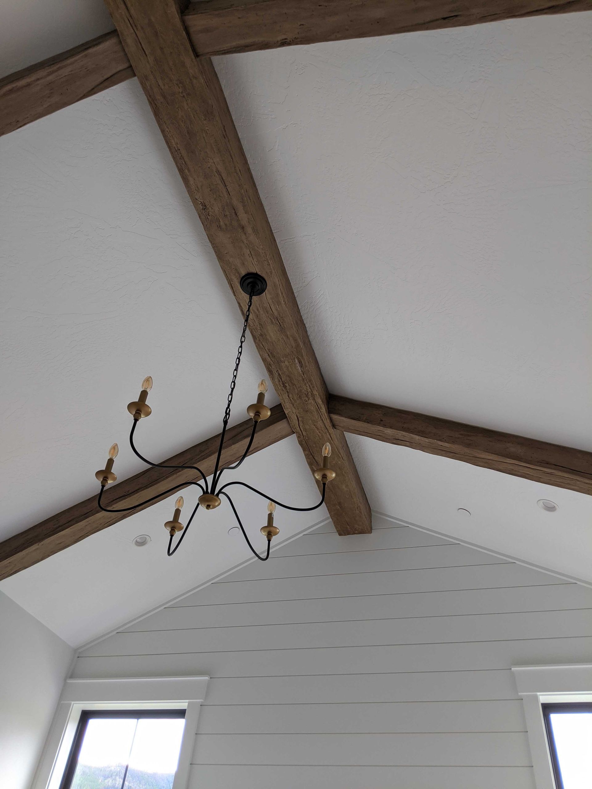 A chandelier hangs from a vaulted ceiling with dark wood beams and white walls, featuring shiplap siding below.