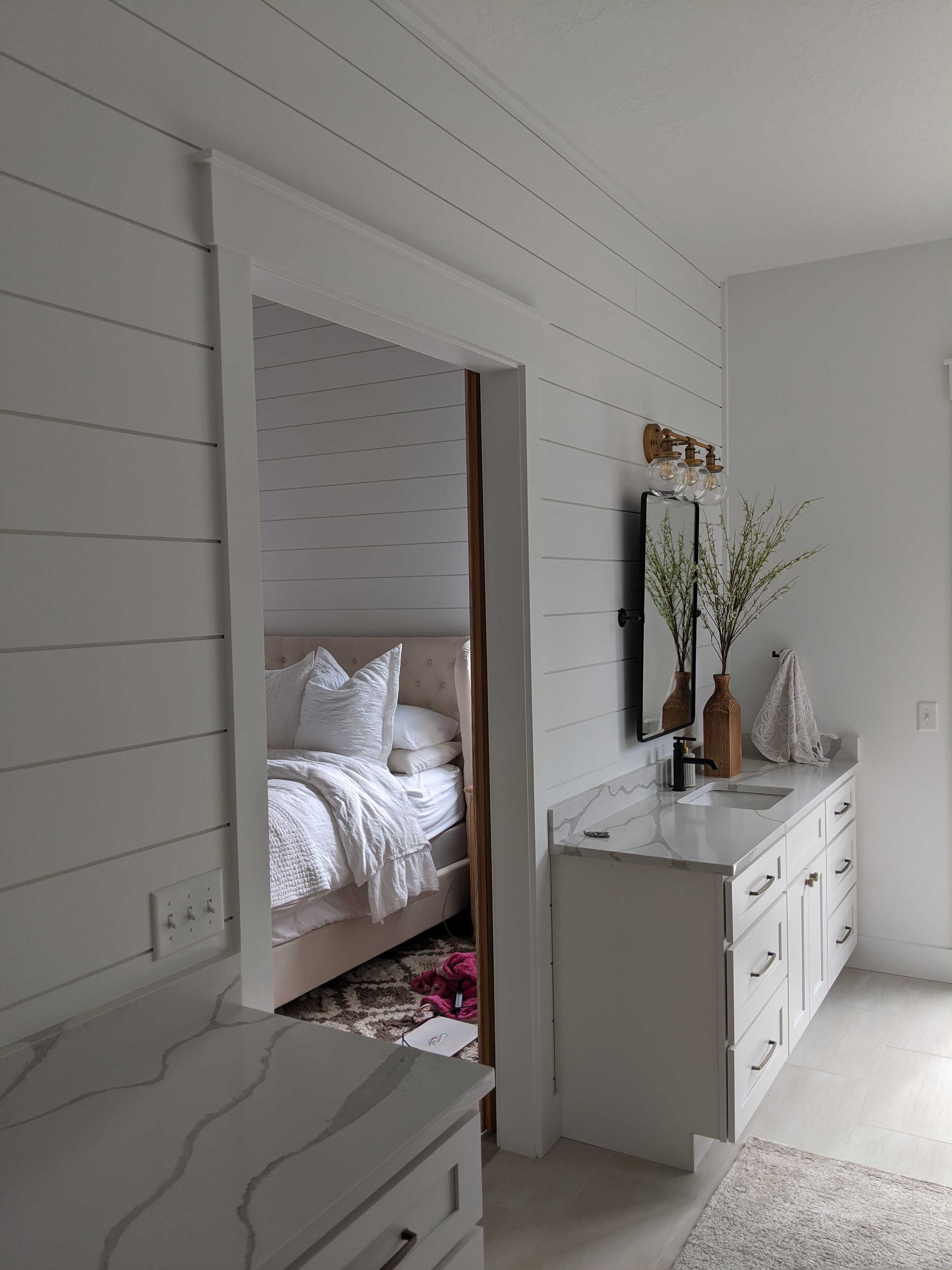 A bright white bathroom with shiplap walls, a marble-topped vanity, and an open doorway looking into a bedroom.