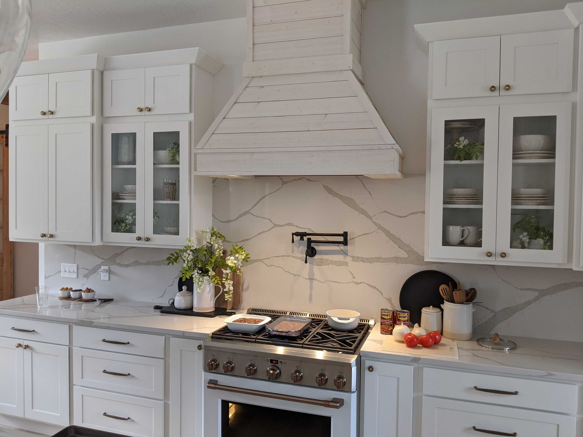 A farmhouse-style kitchen with white cabinets, marble countertops, a wooden range hood, and a stove with a pot filler.