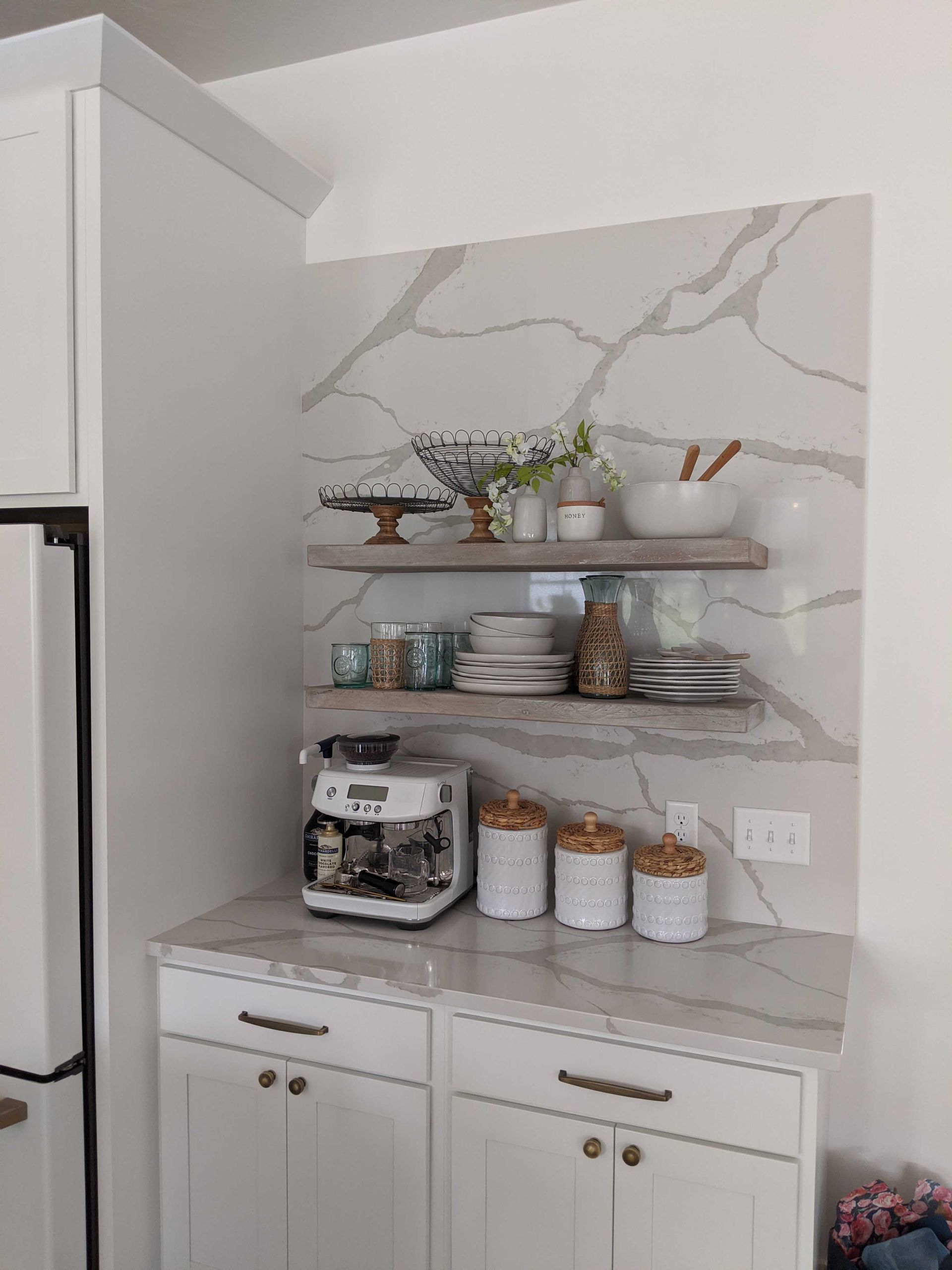 A marble-tiled coffee nook with white cabinets, two wooden floating shelves, a white espresso machine, and canisters.