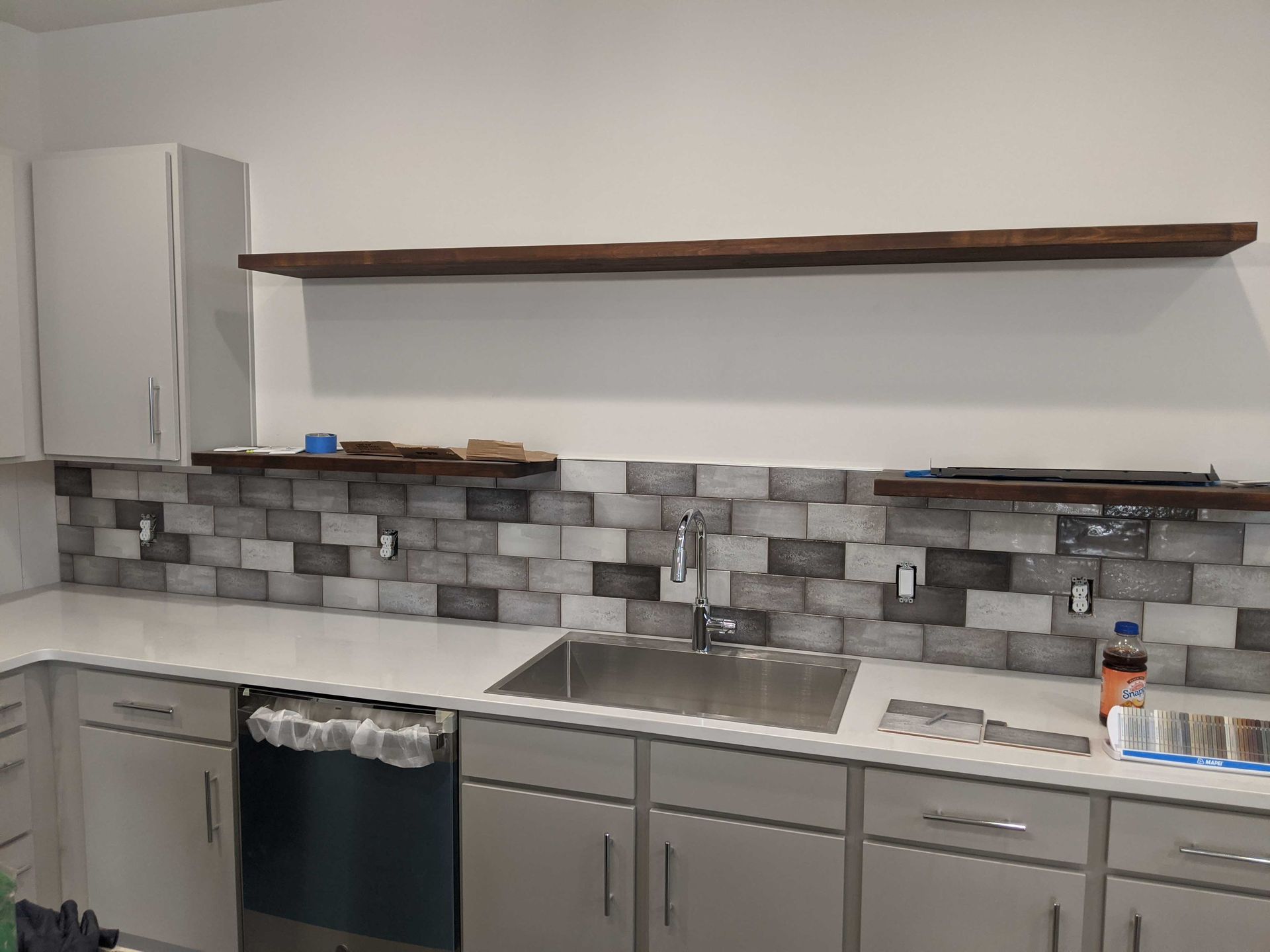 A modern kitchen features gray cabinets, white countertops, a gray tiled backsplash, and floating wooden shelves.