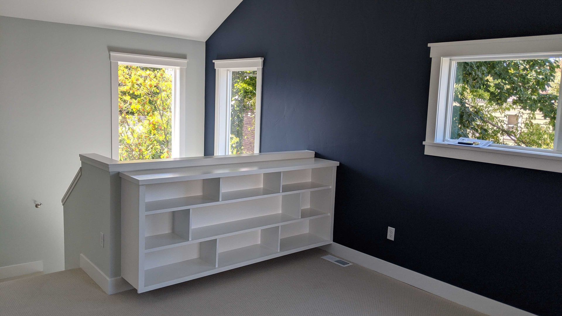 A white bookshelf sits below windows in a room with a light gray wall and a contrasting dark navy blue wall.