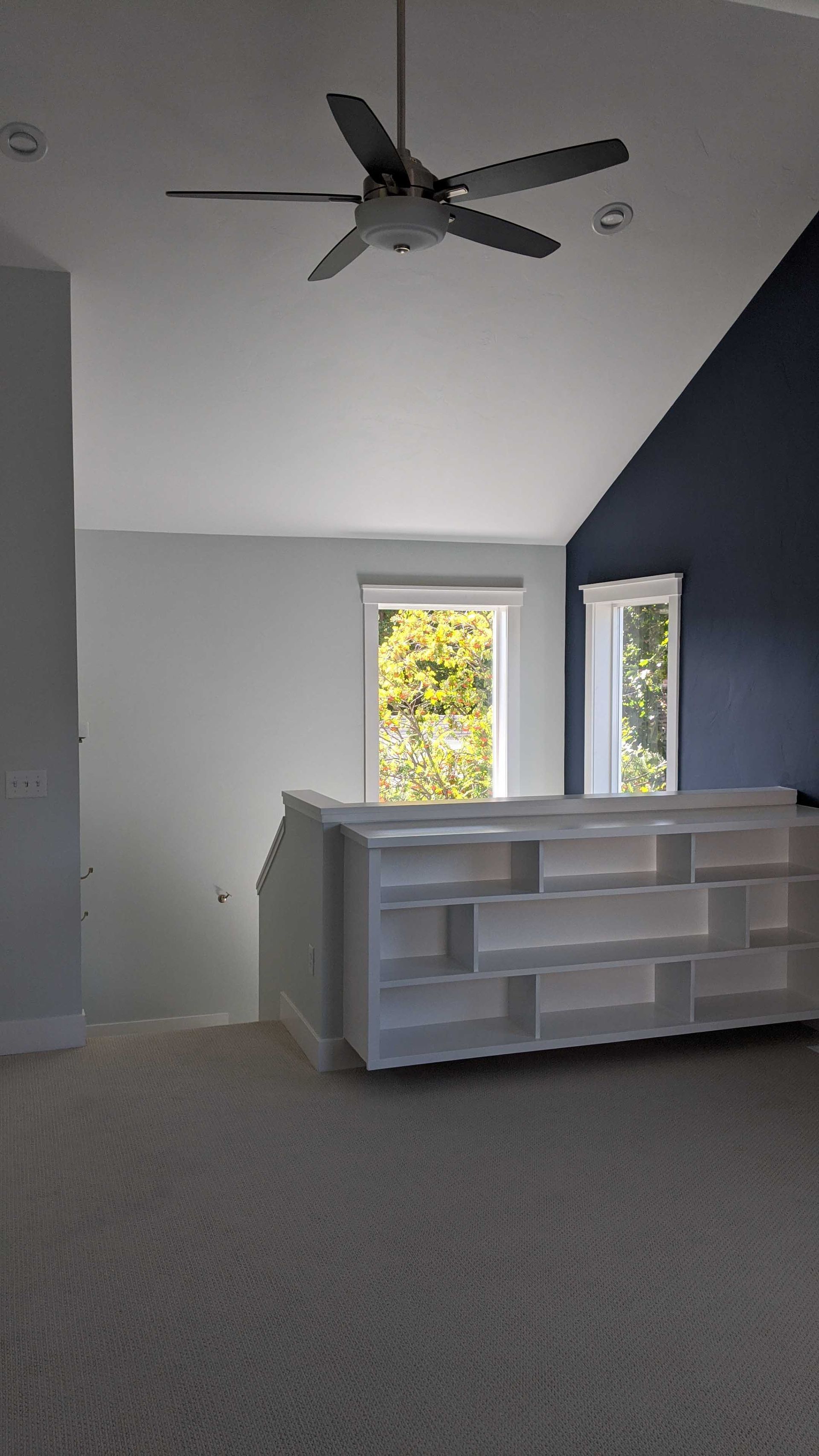 A modern loft area with a low white shelving unit, a ceiling fan, light walls, one dark navy accent wall, and two windows.