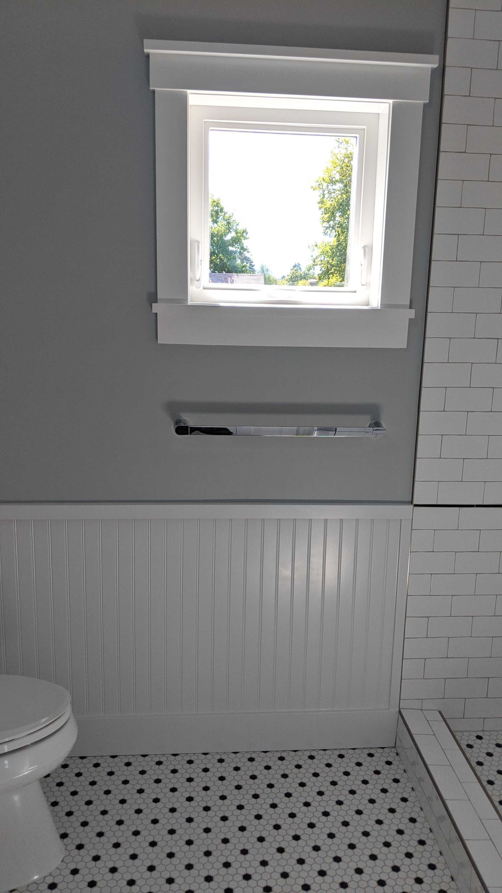 Bathroom view with a window, a towel bar on a grey wall, white wainscoting, and a black-and-white tile floor.