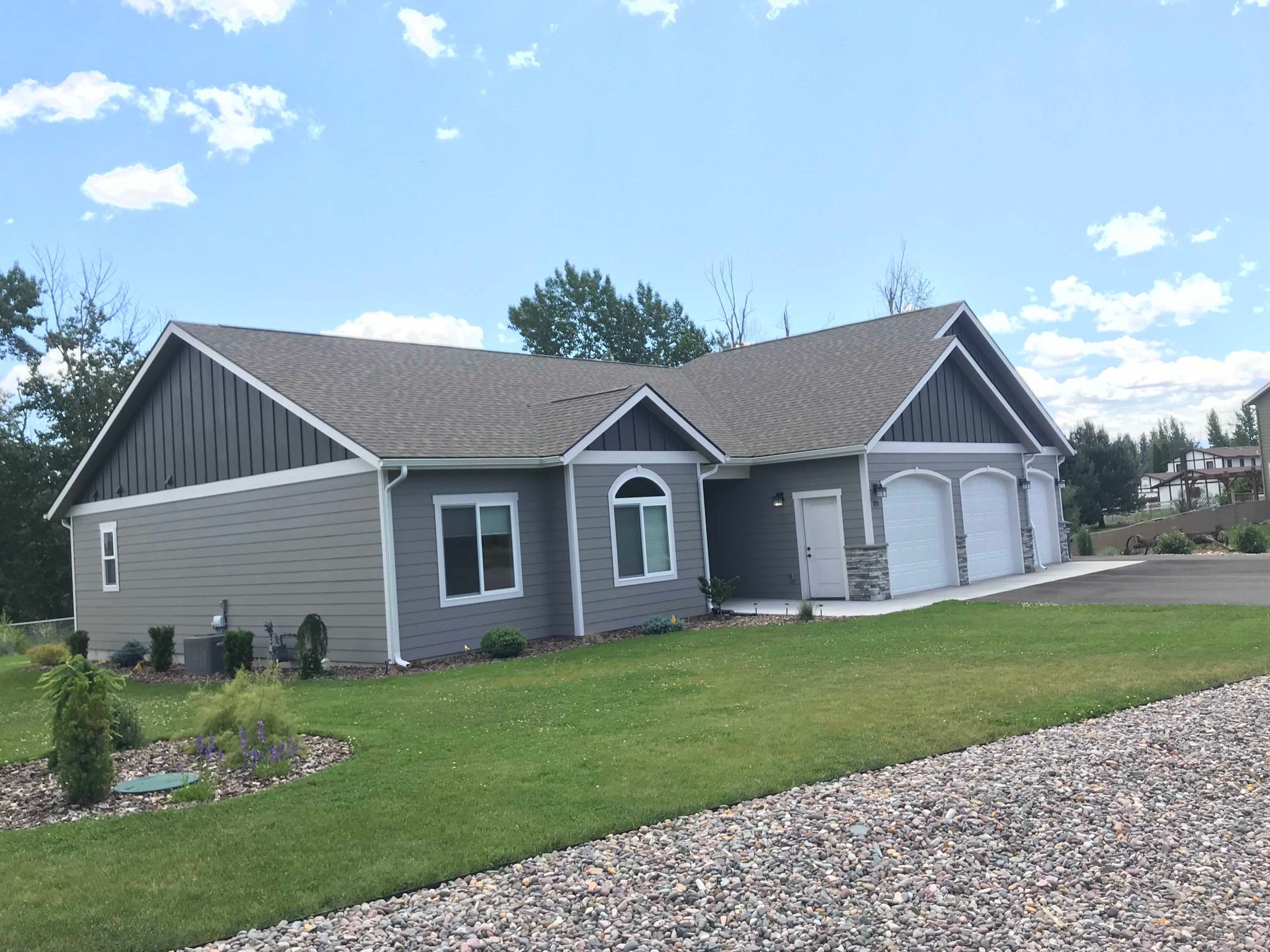 A modern, gray, single-story house with a multi-car garage, a gray shingled roof, and a green lawn under a blue sky.
