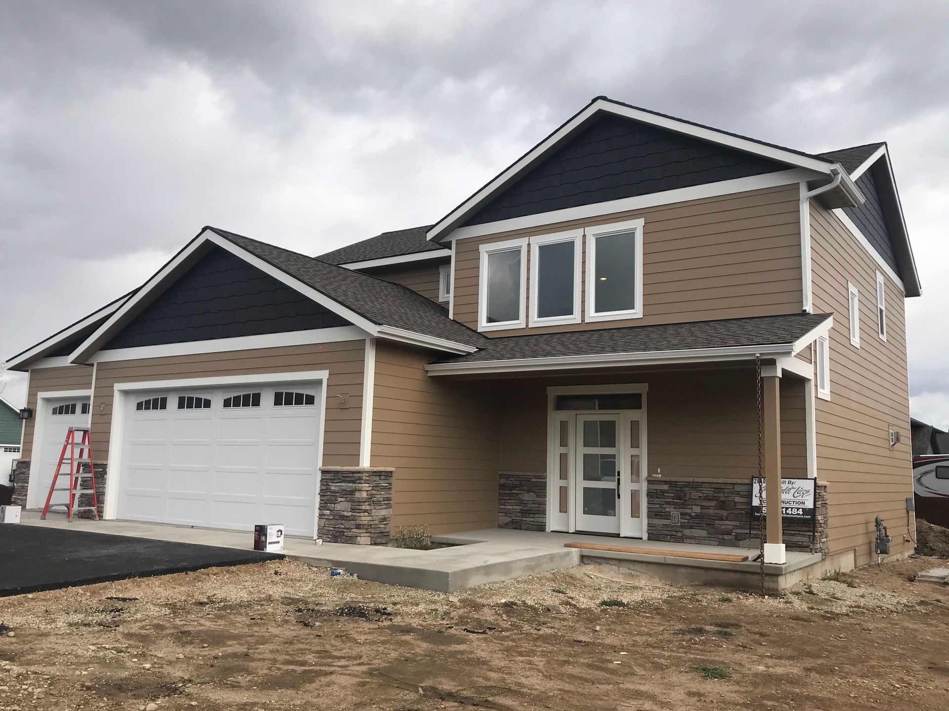 A two-story suburban home under construction with tan siding, blue gables, a stone base, and a white garage door.