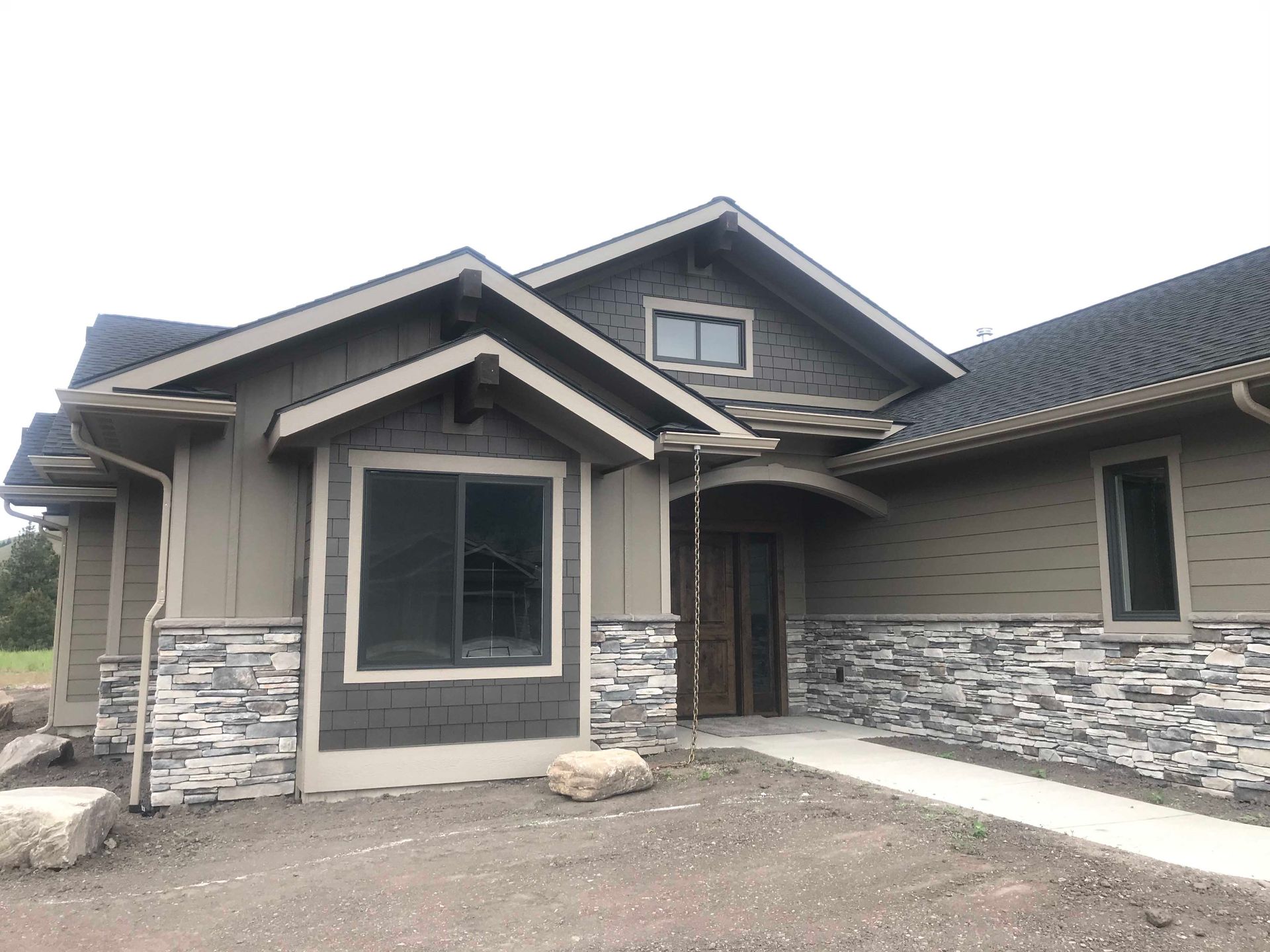 A single-story suburban house exterior with tan siding, stone veneer accents, a dark shingled roof, and a front entryway.