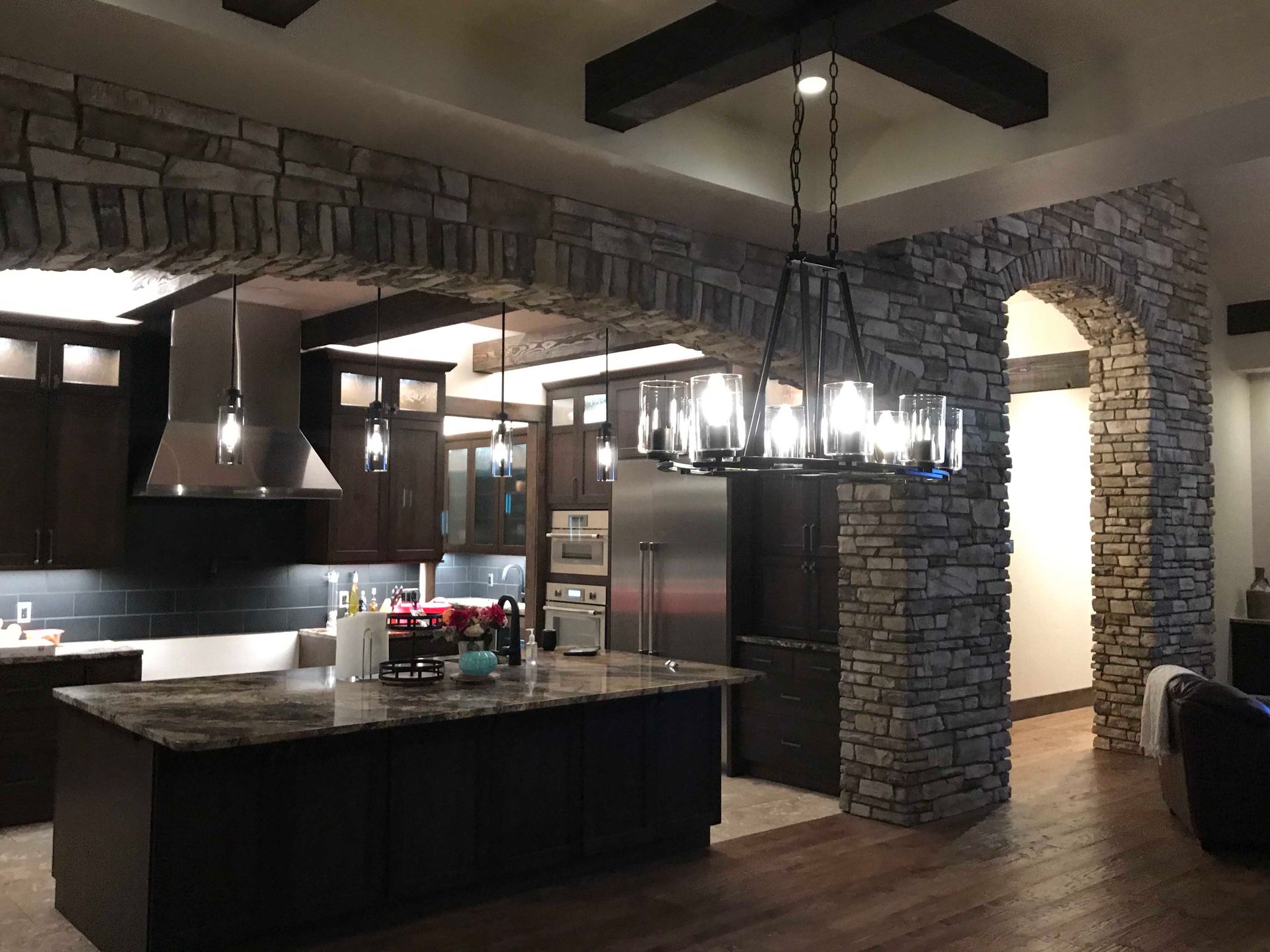 A rustic kitchen with dark cabinetry, granite island, stone arches, and a large metal chandelier hanging from wood beams.