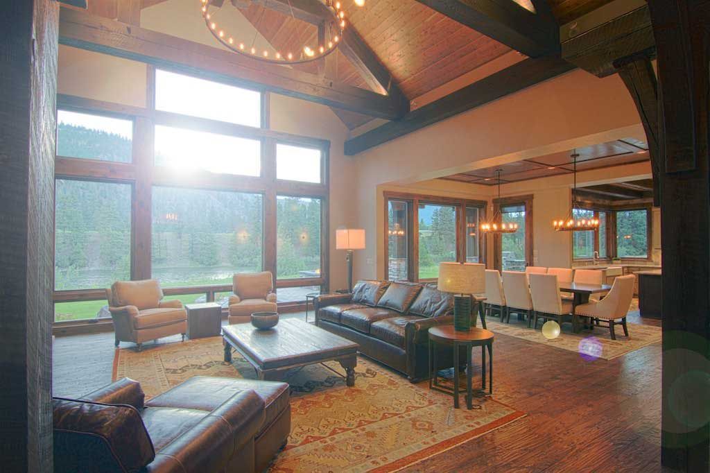 A rustic living room with exposed wood beams, a large window view, leather furniture, and an open dining area.