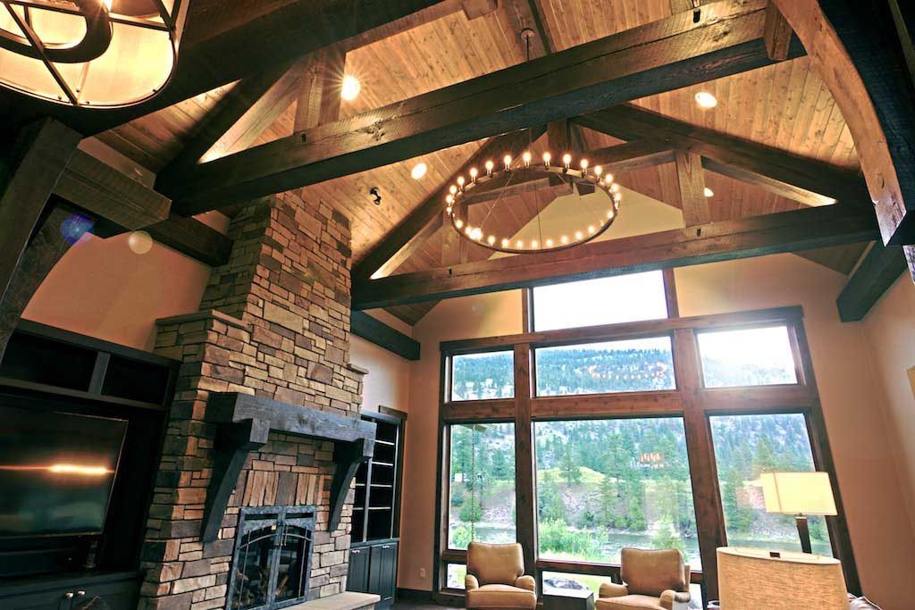 A rustic living room with exposed wood ceiling beams, a stone fireplace, and a large window overlooking a forest.