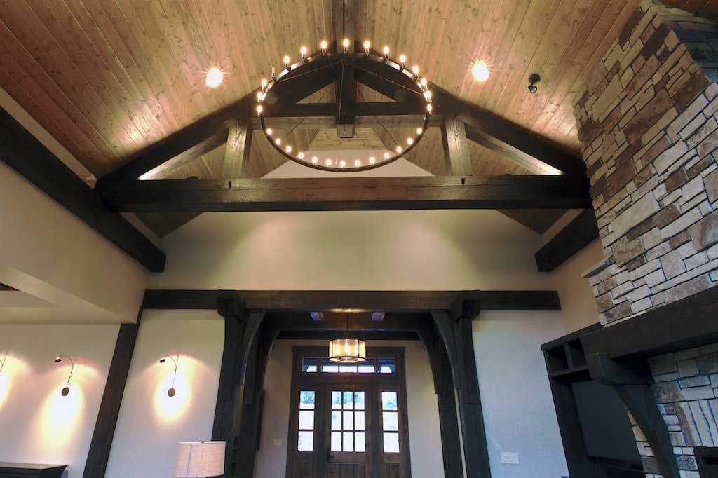 A grand room with wood-paneled vaulted ceilings, exposed dark wooden beams, a circular chandelier, and a stone fireplace.