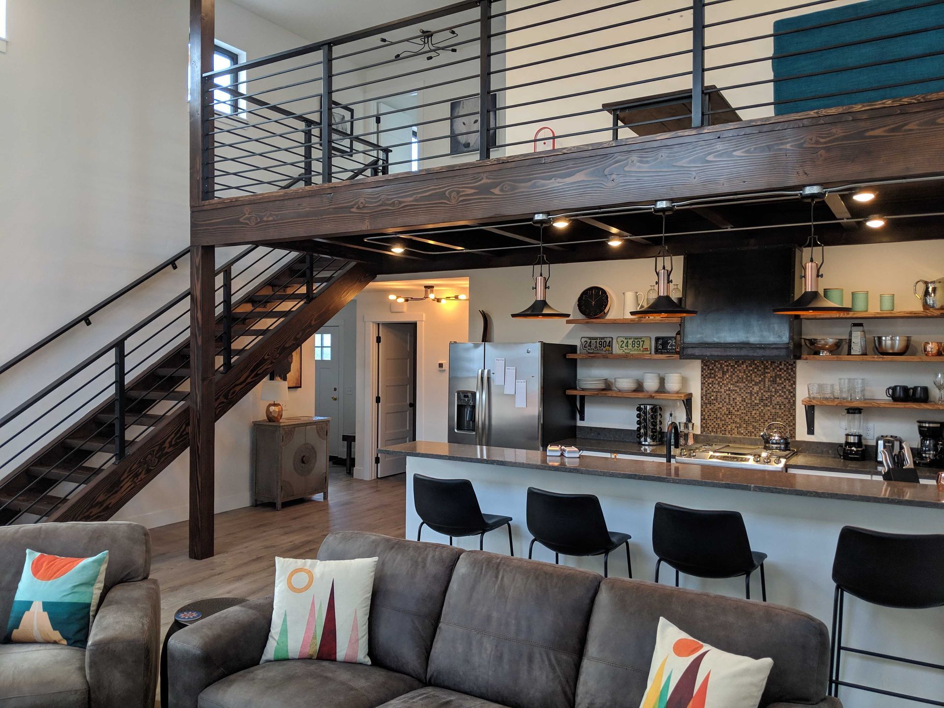 Open-plan living room with a loft area above a modern kitchen, featuring a grey sofa, black bar stools, and stairs.
