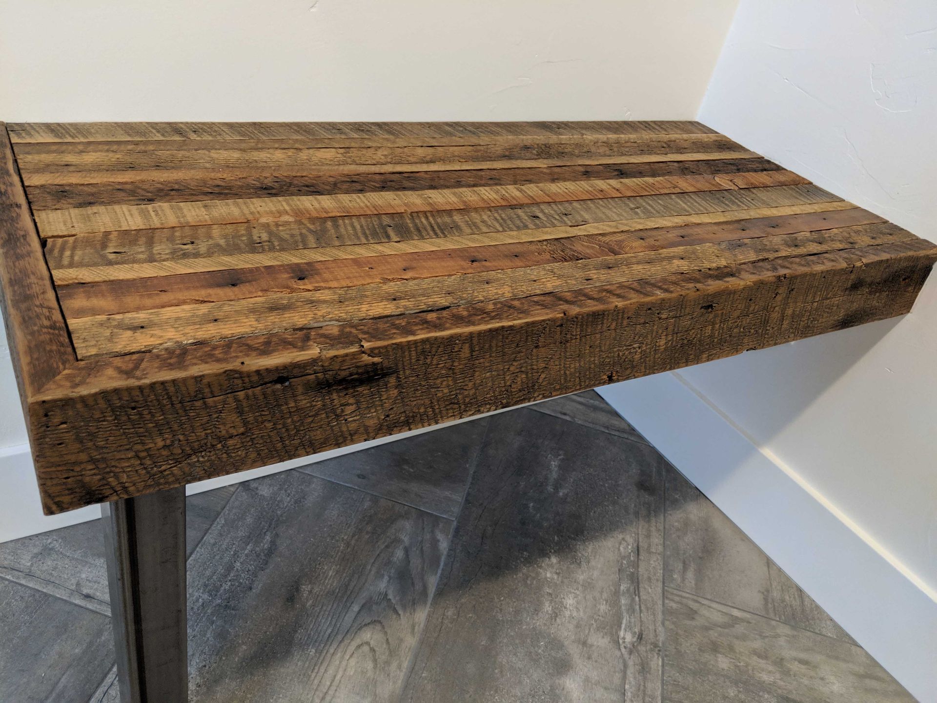 A rustic, rectangular wooden console table with a thick, textured top and a metal leg against a white wall.