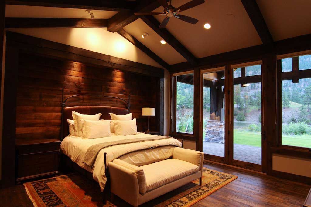 A luxury rustic bedroom with dark wood paneling, a bed, a bench, and floor-to-ceiling windows overlooking a green landscape.