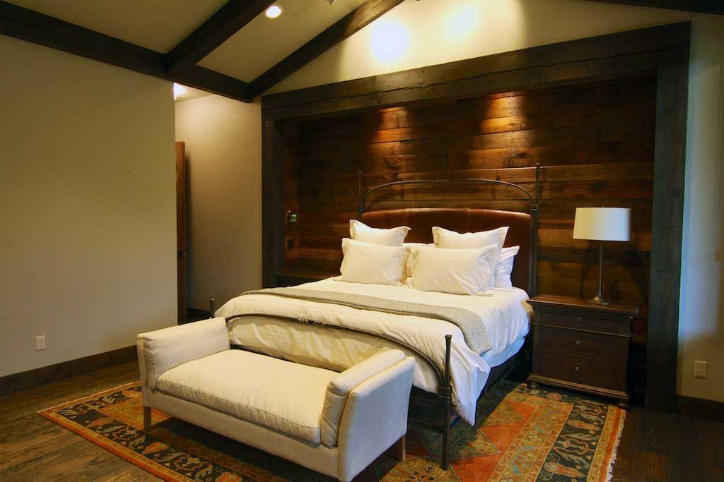 A rustic bedroom with a wooden paneled wall, vaulted ceiling, white-bedding, and a cream-colored bench on a patterned rug.