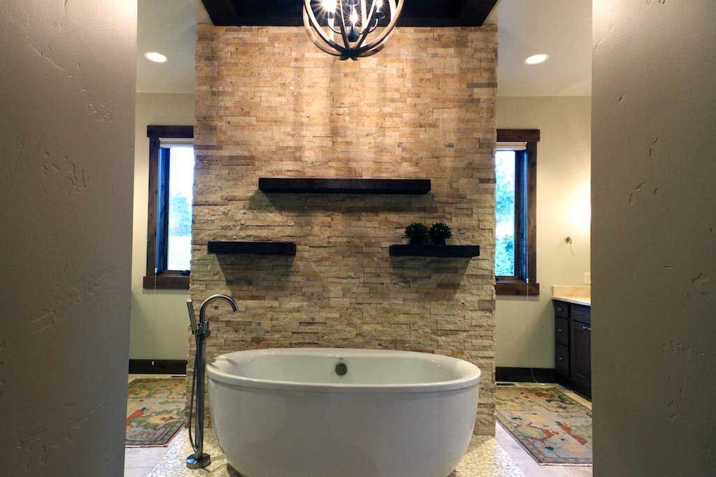 A white freestanding bathtub set against a textured stone wall with three black floating shelves and a chandelier above.