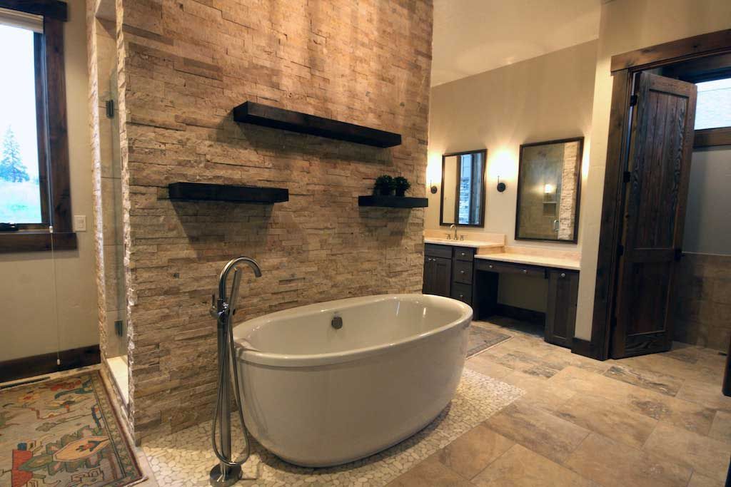A freestanding white bathtub sits against a stone wall featuring three black floating shelves in a modern bathroom.