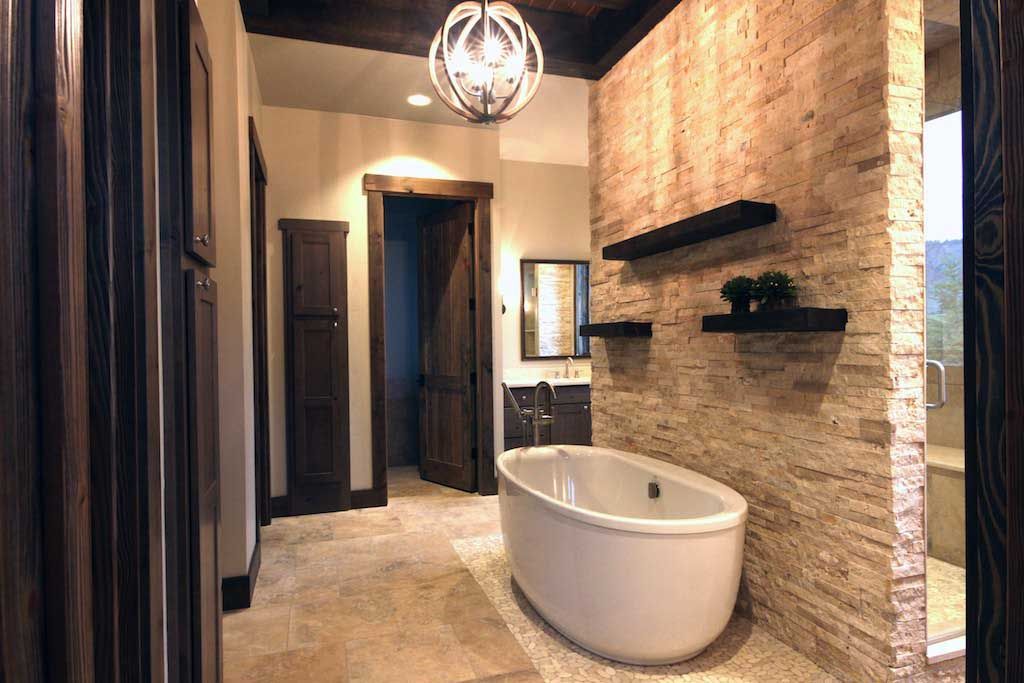 A modern bathroom featuring a white soaking tub against a textured stone wall with dark wood trim and shelving.