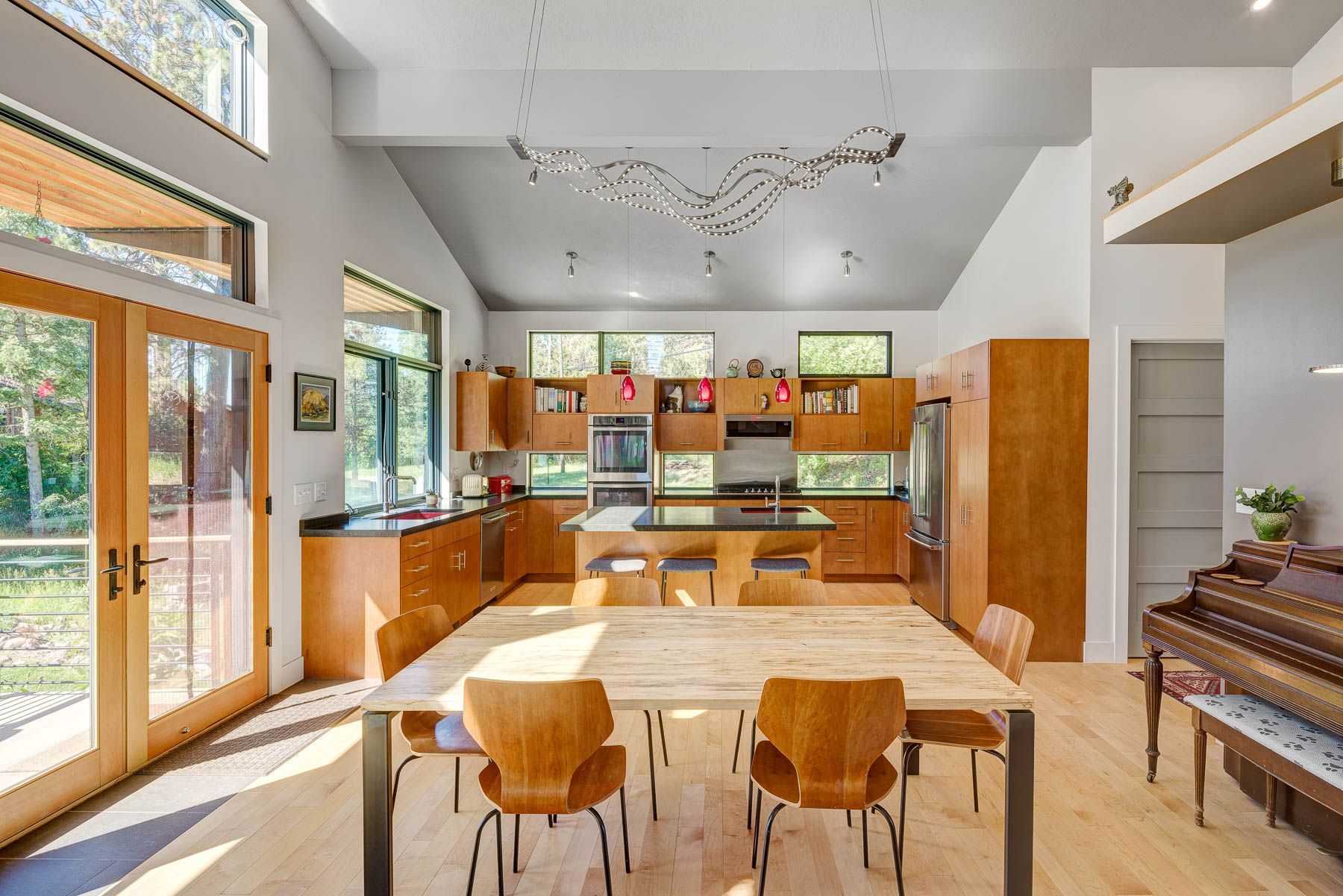 A bright, open-plan kitchen and dining area with wooden cabinets, a light wood dining table, and a piano.