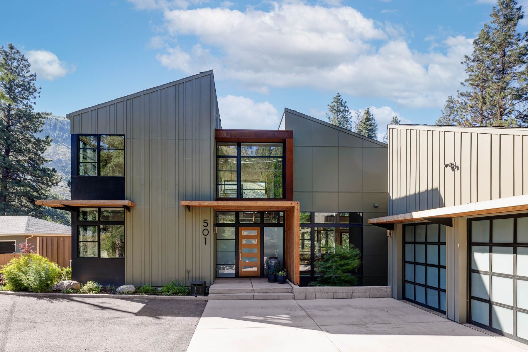 A modern two-story home with olive-green metal siding, large windows, an entryway, and a two-car garage under a blue sky.