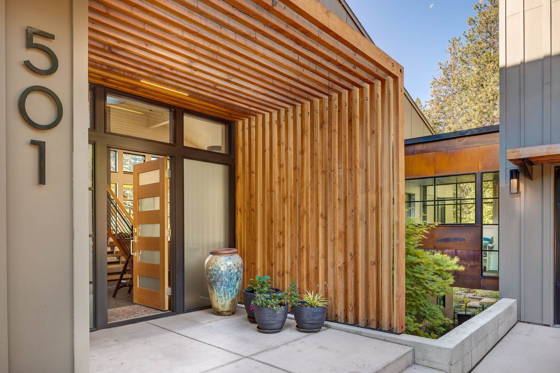 Modern home entrance featuring a wood-slat portico, glass panel door, house number 501, and potted plants on a concrete pad.
