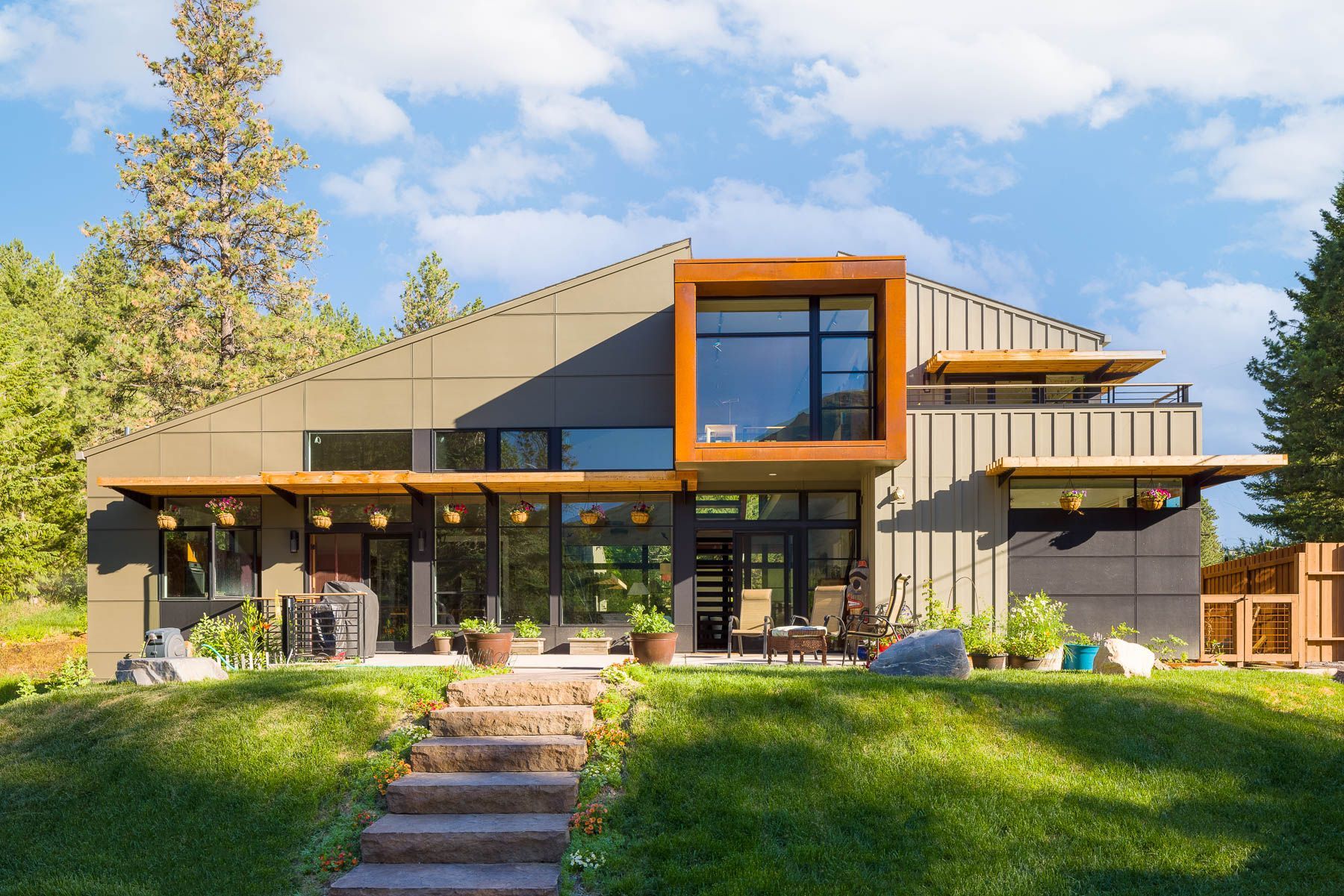 A modern house with slanted rooflines, wood accents, and large windows sits on a grassy hill with stone steps.