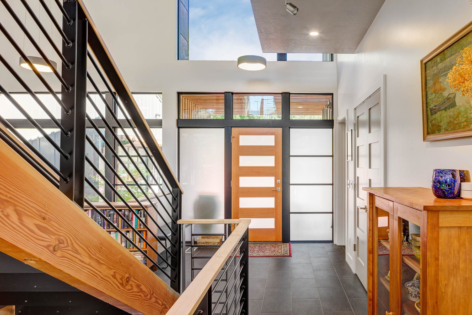 A modern entryway featuring a wooden door, a staircase with black metal railings, a tiled floor, and a wooden console table.