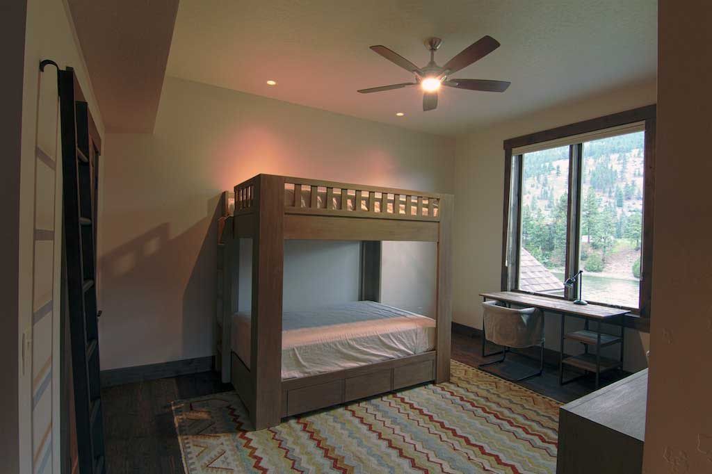 A bedroom with a wooden bunk bed, a desk by a large window, a patterned rug, and a ceiling fan.