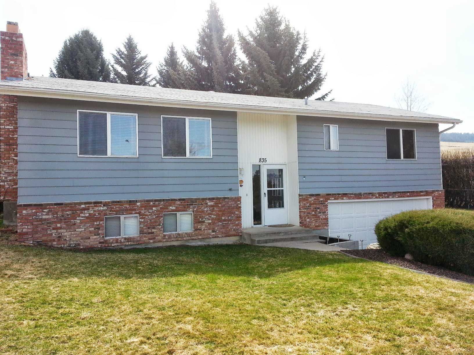 Split-level house with blue siding, a brick base, white trim, and a one-car garage, surrounded by a lawn and tall trees.