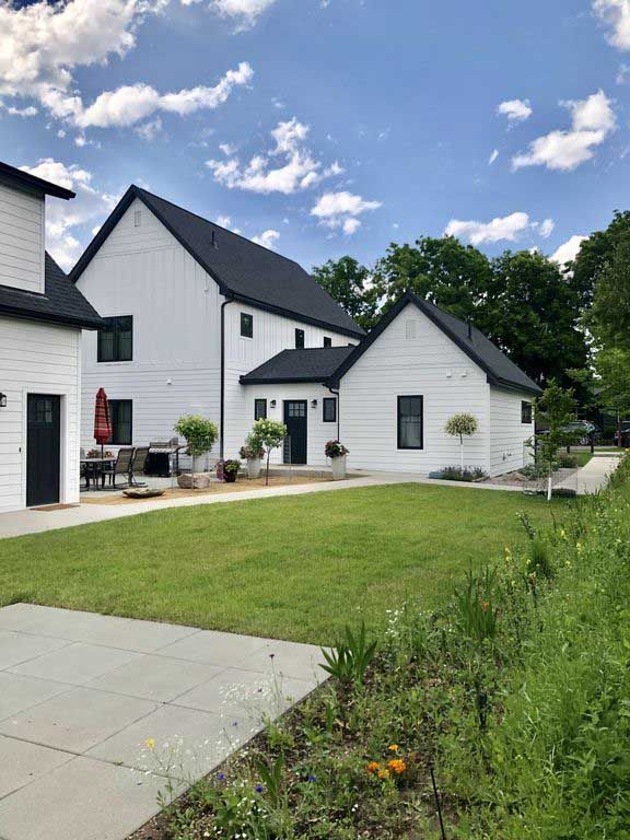 A white modern farmhouse with a black roof and matching trim, surrounded by a lawn and landscaped garden under a blue sky.