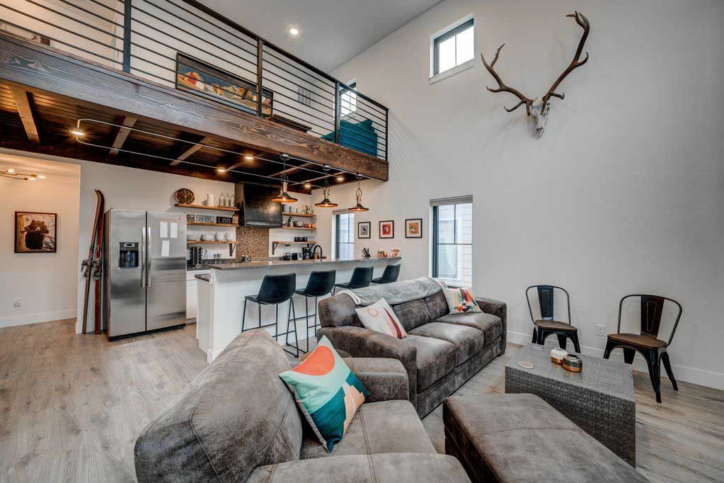 A rustic, modern living room featuring a gray sofa, a large antler wall mount, and an open loft area with a kitchen below.