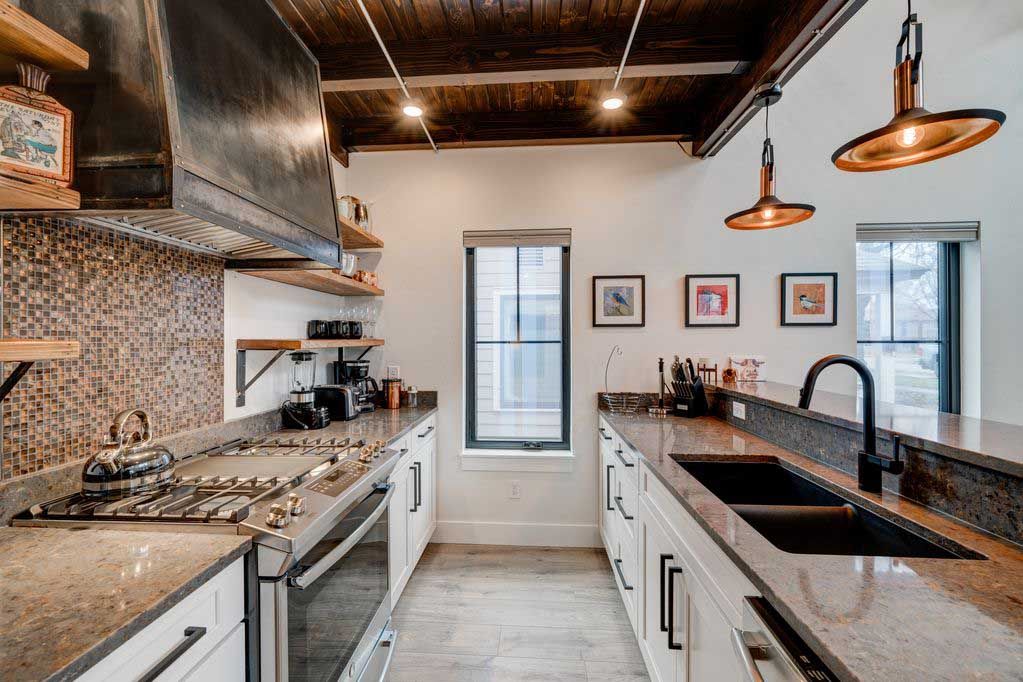 A galley-style kitchen with white cabinets, speckled countertops, a dark vent hood, wood ceiling, and two pendant lights.