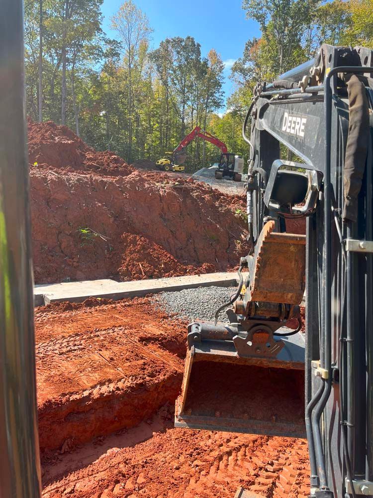 A view from inside an excavator cab showing a construction site with red dirt, gravel, and a distant crane.