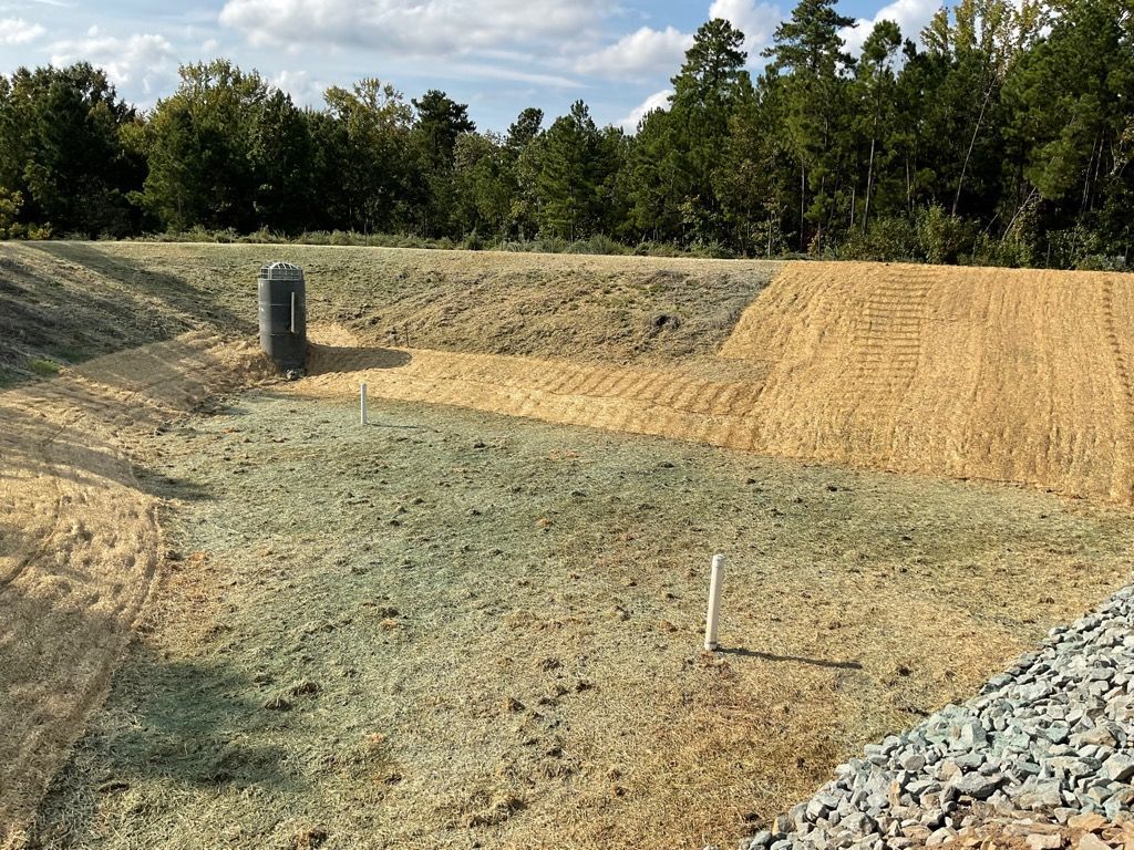 A newly graded stormwater retention basin featuring straw erosion control matting, two vertical pipes, and a stone spillway.