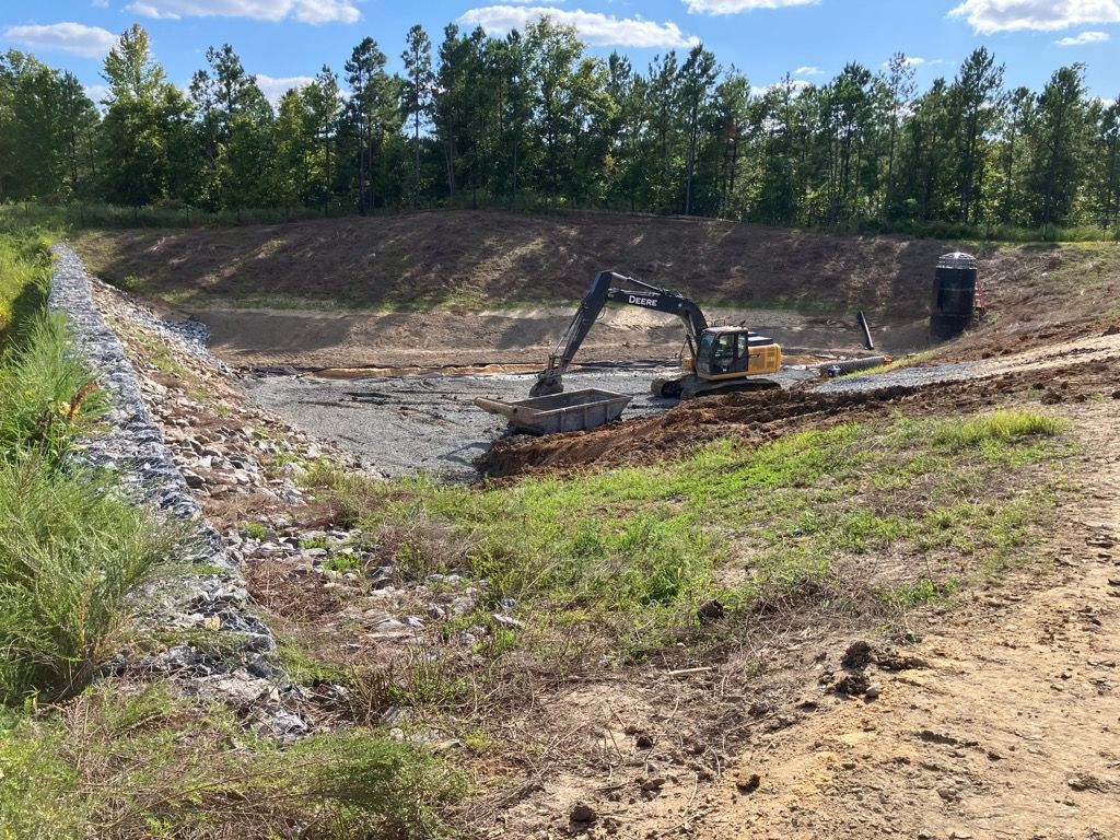 A yellow excavator works in a dirt clearing bordered by a rock-lined slope and trees under a blue, cloudy sky.