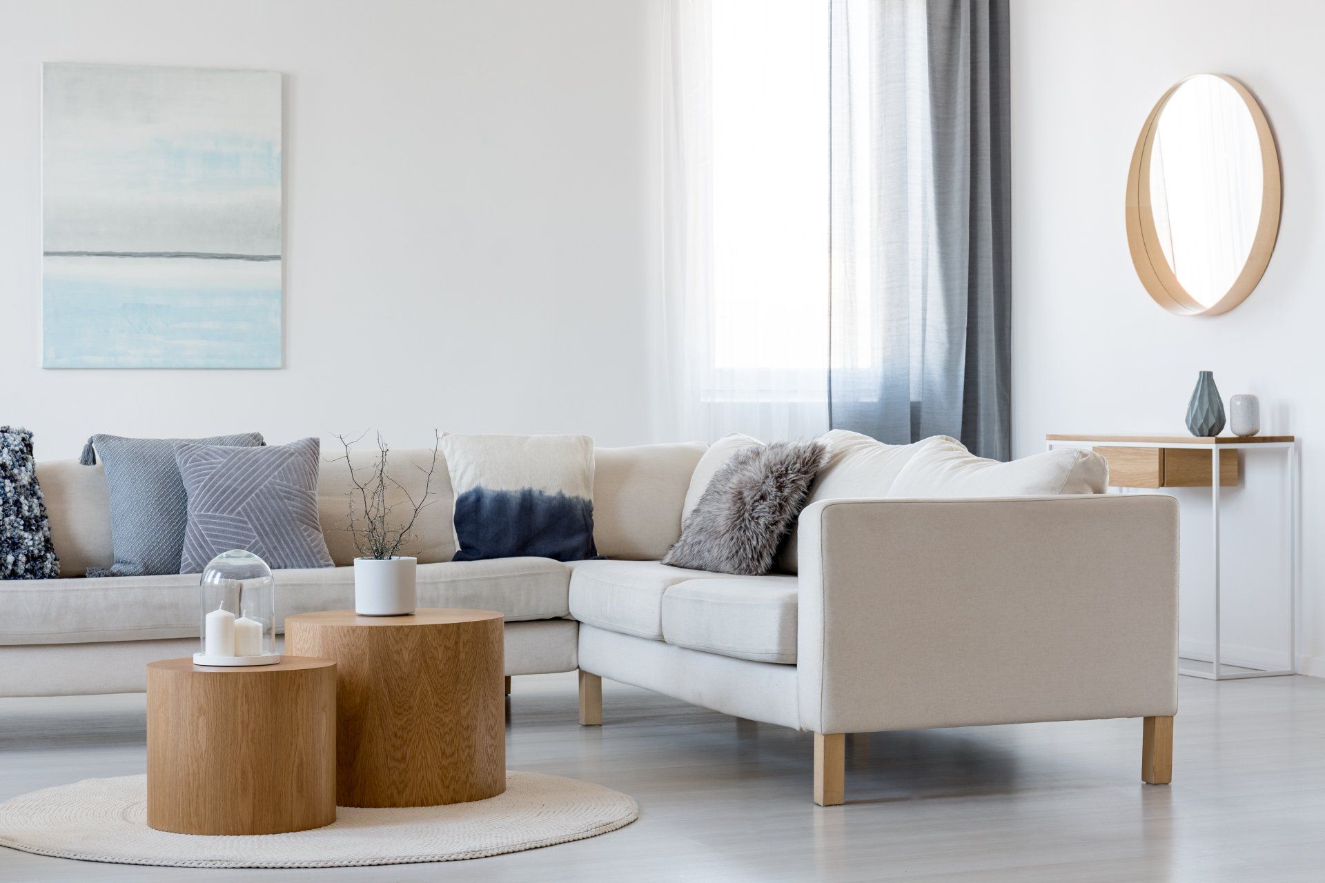 L-shaped beige sofa in a white living room with light blue decor, wood tables, and a round mirror.