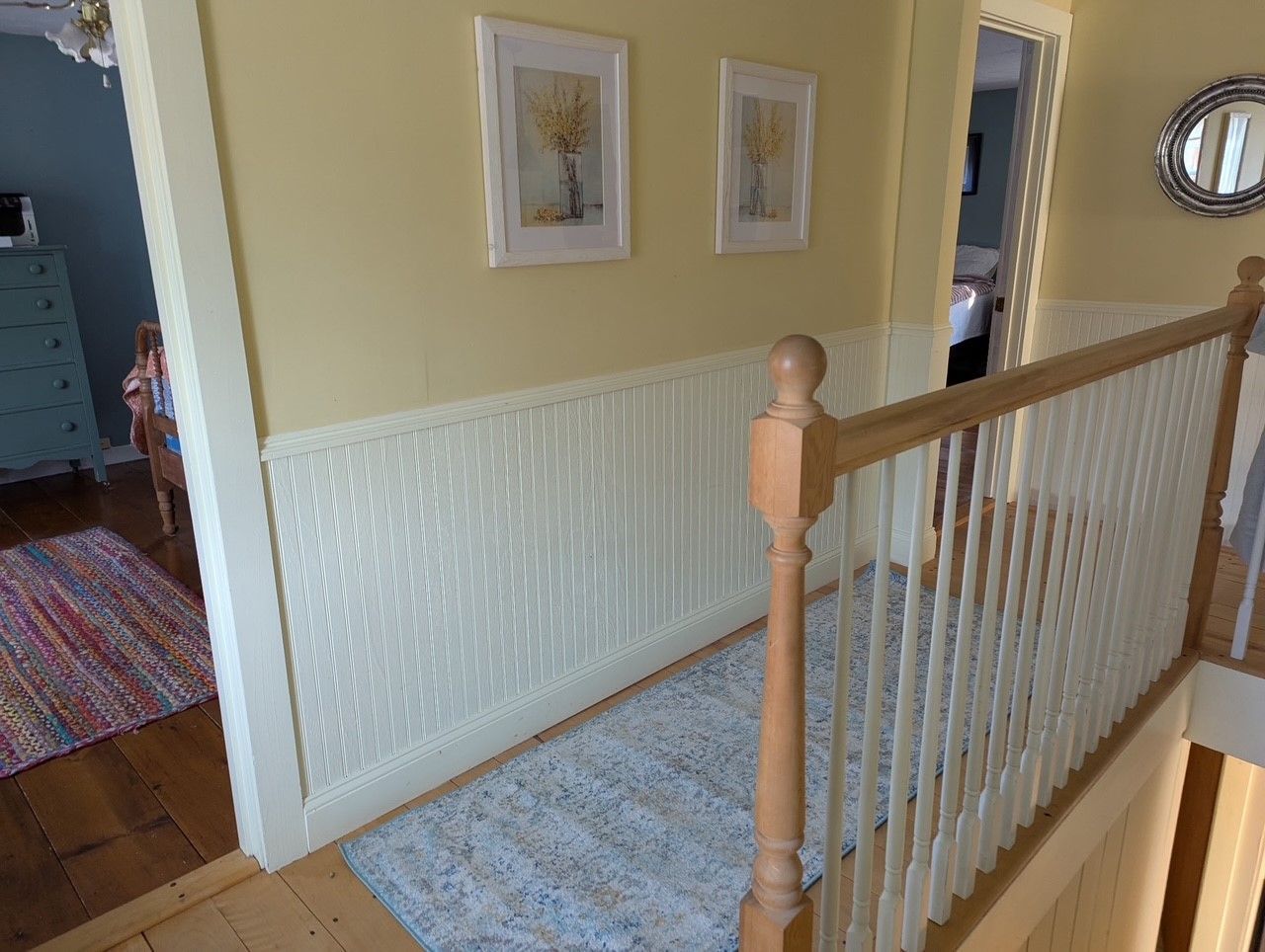Hallway with yellow walls, white wainscoting, and two framed pictures. A railing is on the right.
