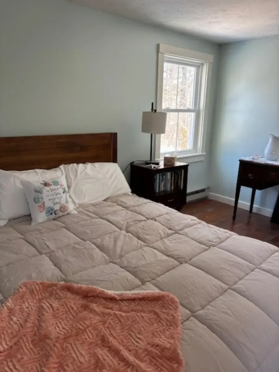 Bedroom with bed, nightstand with lamp, and window with natural light. Soft colors, cozy ambiance.