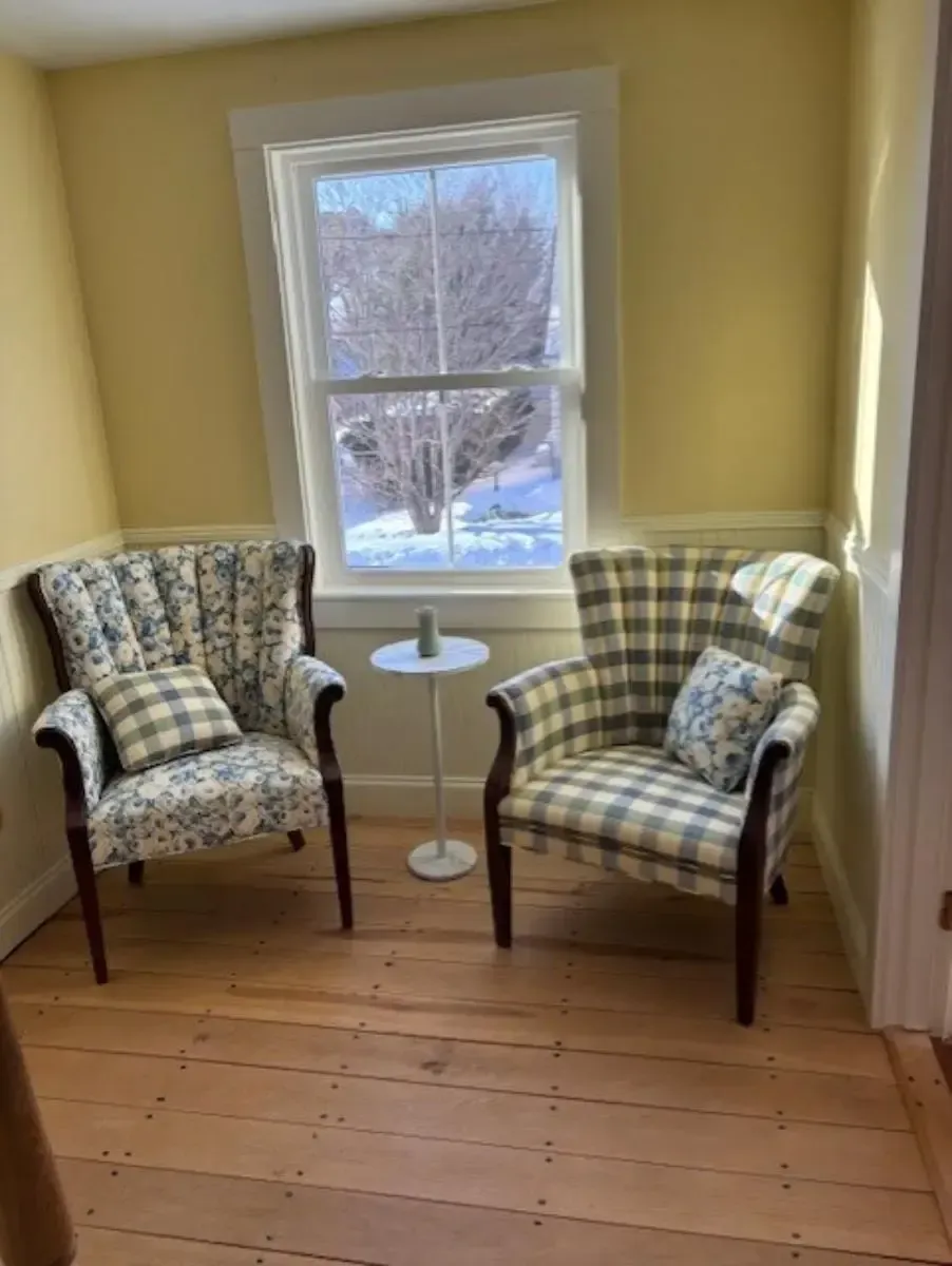 Two armchairs with patterned fabric face a window. A small table sits between them in a yellow room.