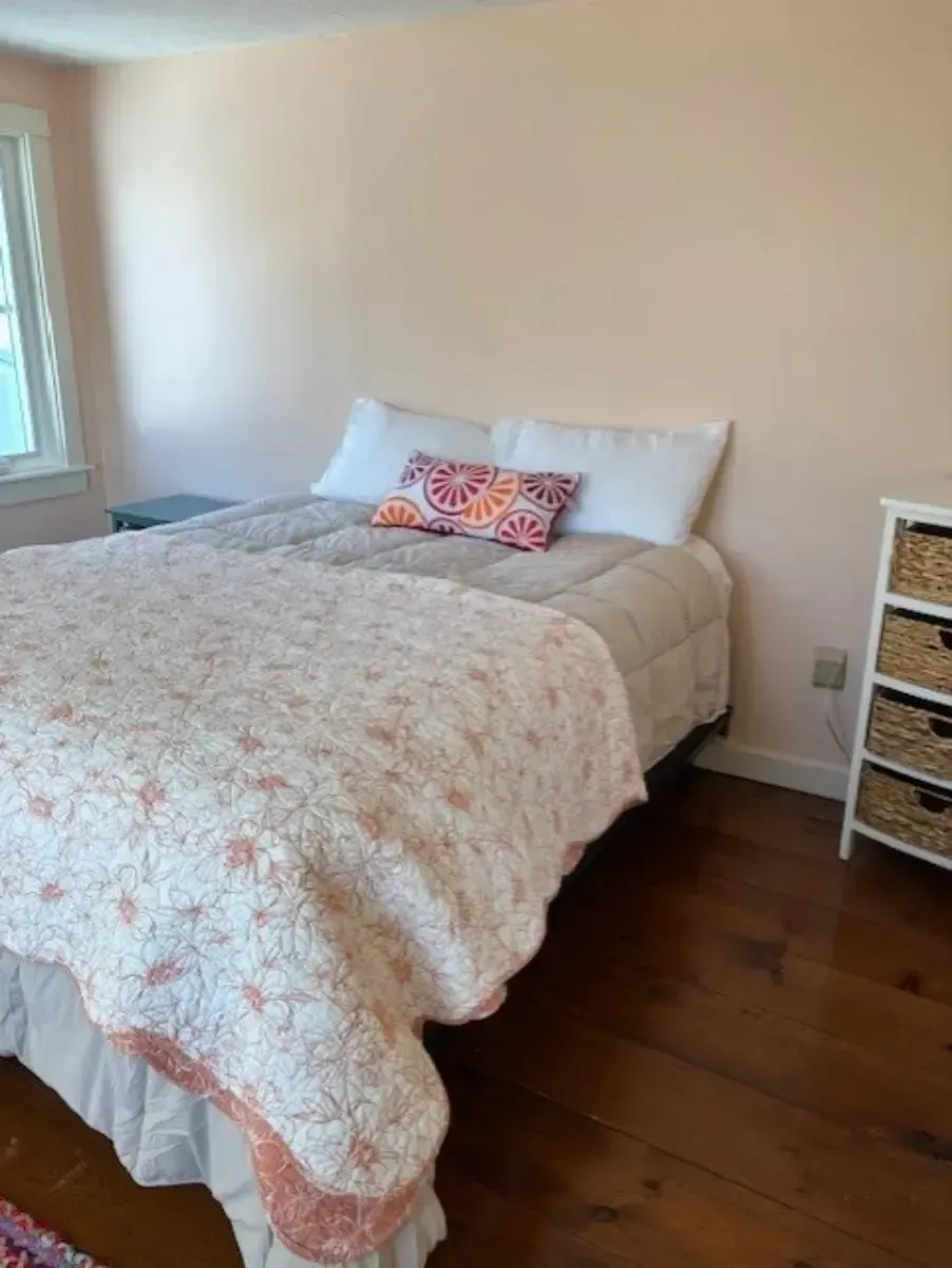 Bedroom with bed covered in a floral quilt, light peach walls, small chest of drawers, and hardwood floors.