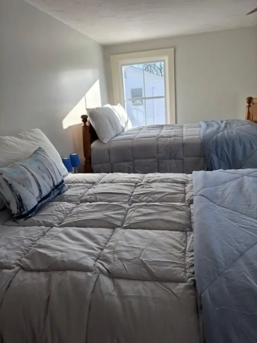 Two beds with gray and blue bedding in a bright room with a window.
