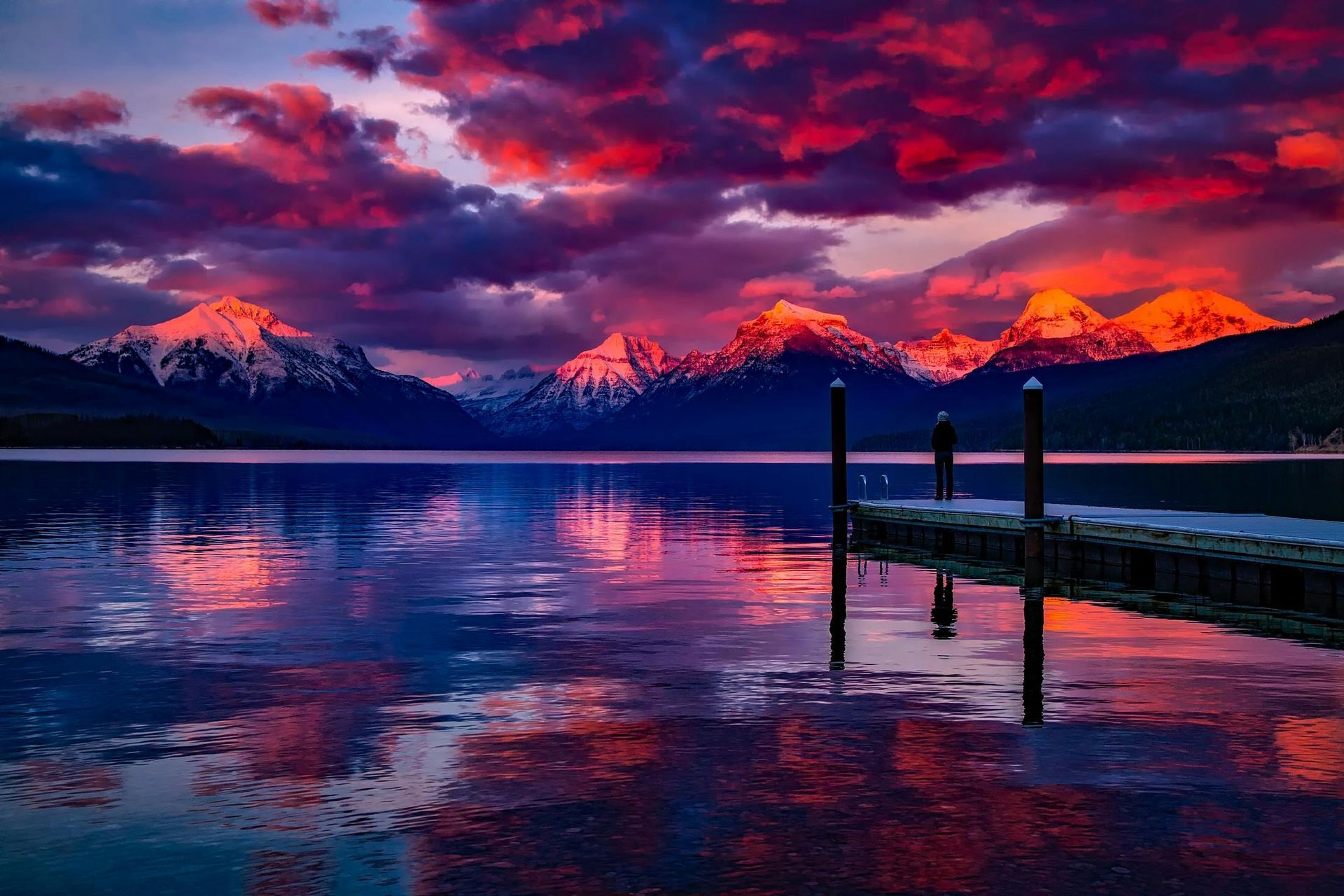 Sunset over snow-capped mountains and a calm lake with a wooden pier, vibrant red and purple sky.