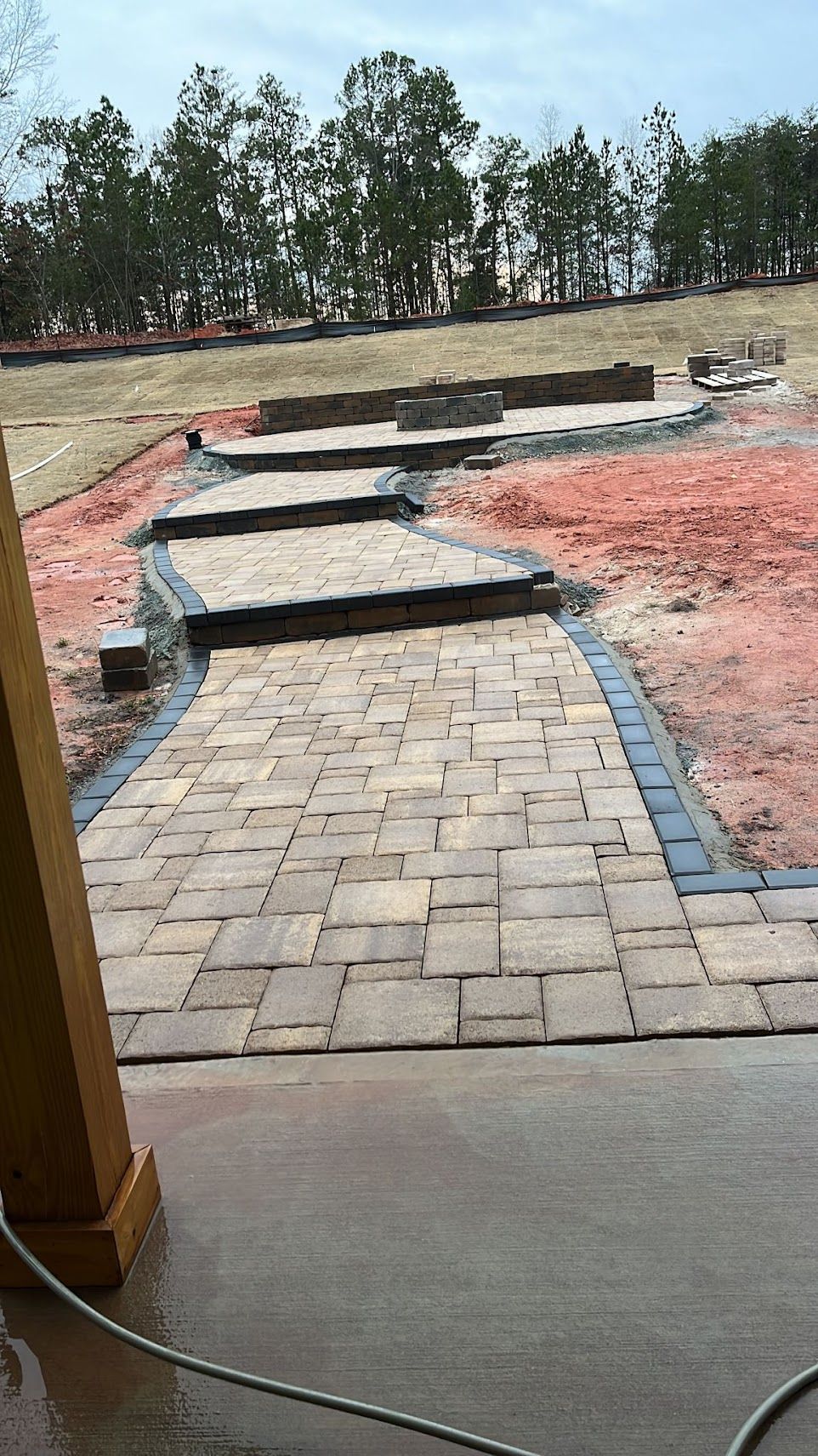 Brick pathway with stone steps leading to a raised area. Soil and trees in background.
