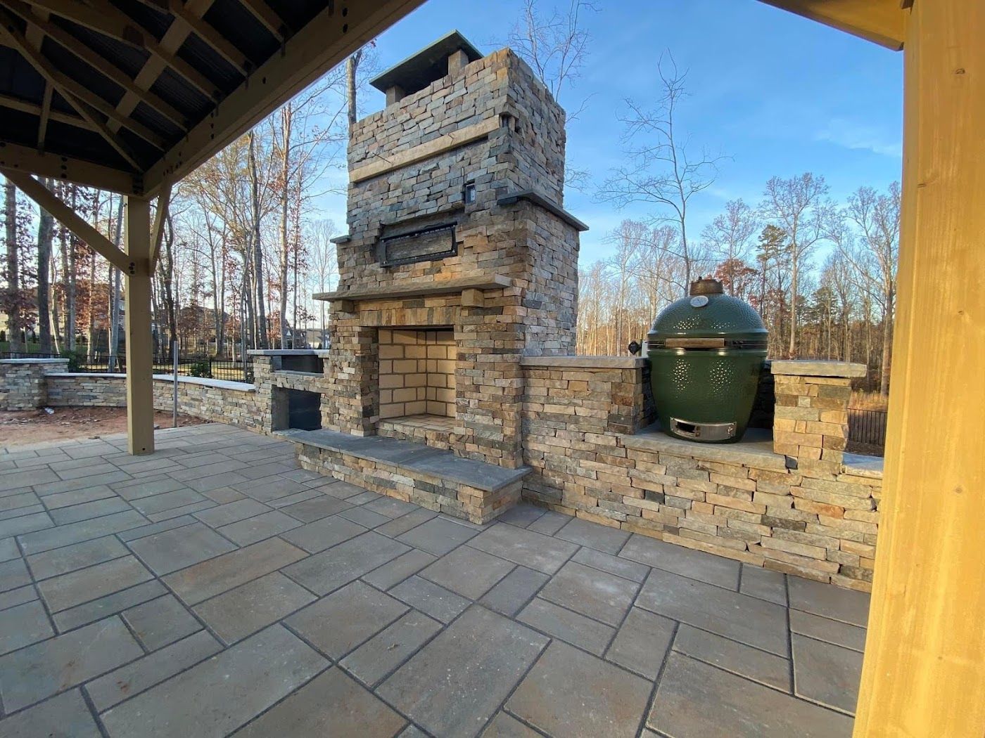 Stone outdoor fireplace with a Big Green Egg grill on a patio under a wooden structure.
