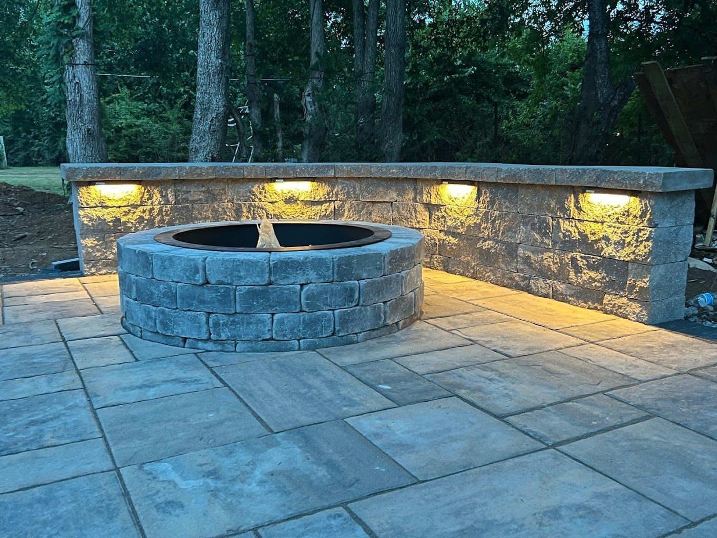 Fire pit on a stone patio with a stone wall lit by warm lights. Trees in background.