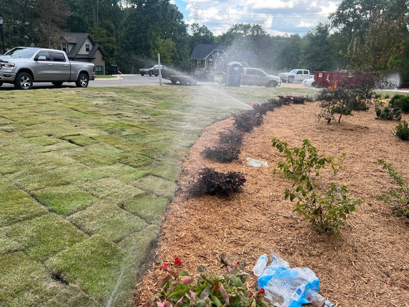 Sprinkler system watering newly laid sod and mulch border in a residential yard. A truck is parked nearby.