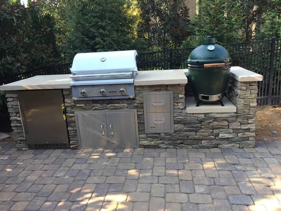 Outdoor stone kitchen with a grill, Big Green Egg, refrigerator, and storage on a paver patio.