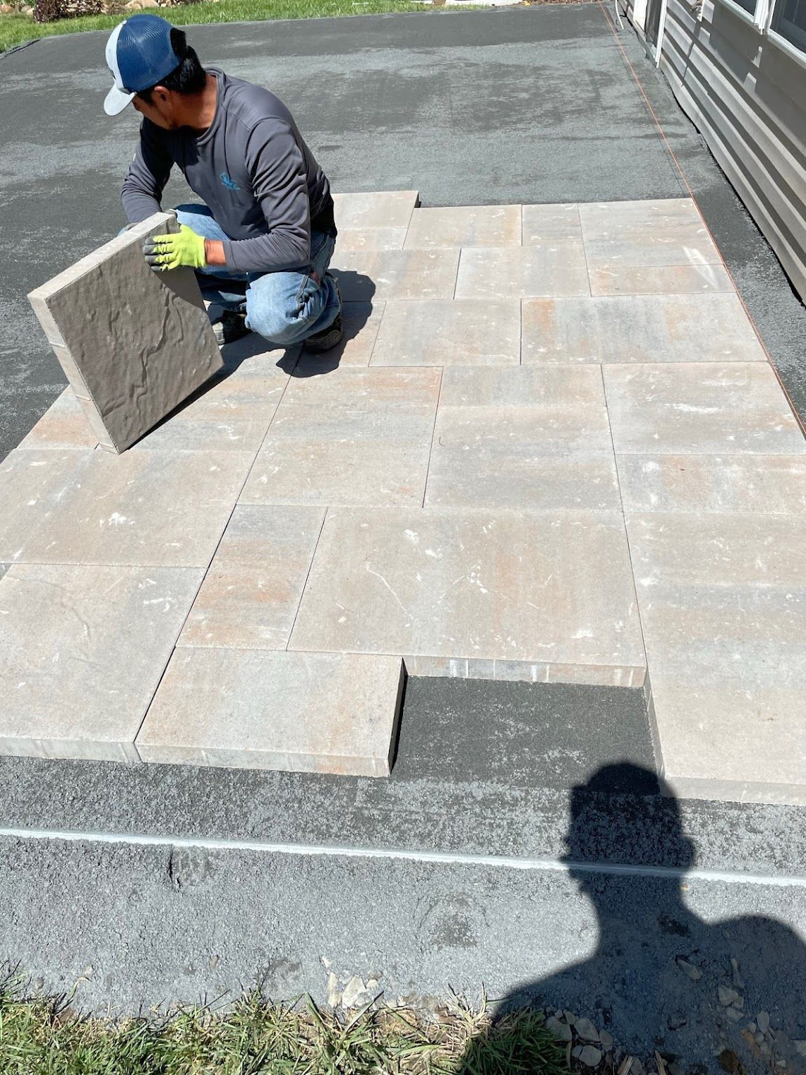 Man laying paving stones on a dark surface, wearing gloves and a cap; sunlight.