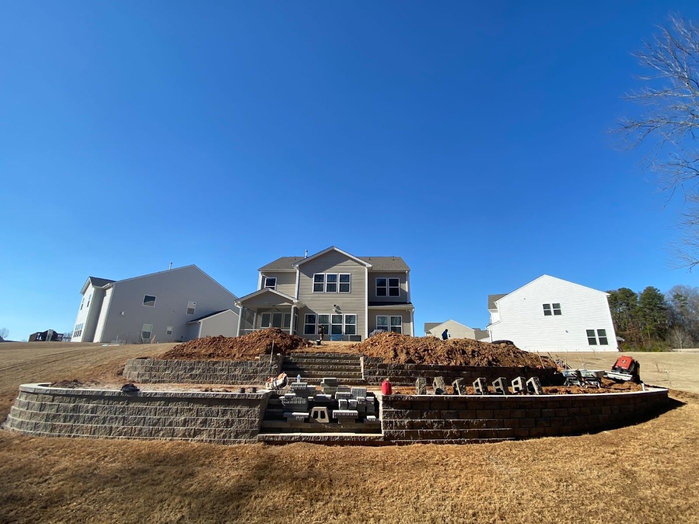 Stone retaining walls and stacked wood in front of a multi-story house on a sunny day.