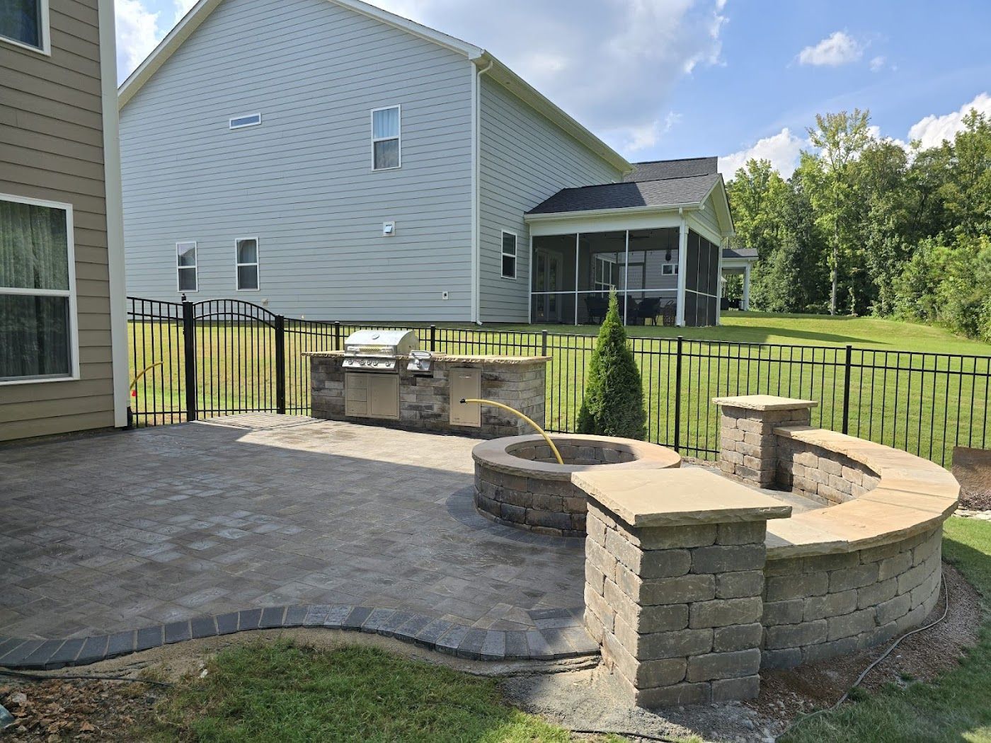 Backyard patio with built-in grill, fire pit, seating, and a house in the background, surrounded by a black fence.